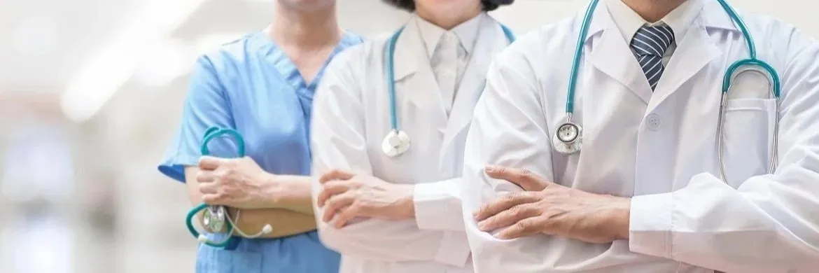 Three clinicians in white coats and scrubs with stethoscopes, arms crossed